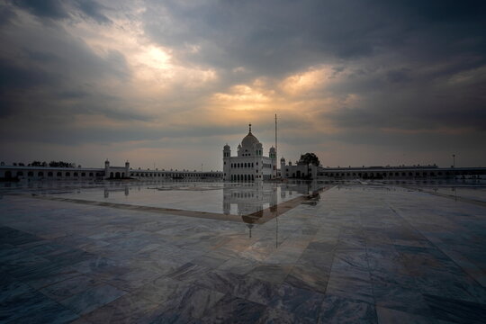 View of Gurdwara Darbar Sahib's serene reflection shimmers across the marble expanse under a dramatic sky, Kartarpur, Punjab, Pakistan.