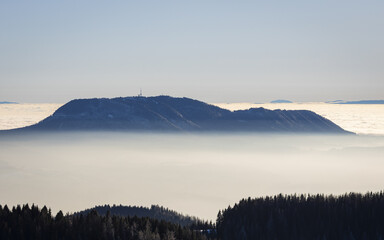 View of the majestic mountain Schöckl shrouded in ethereal mist, with the dark silhouettes of trees contrasting against the bright, hazy sky, Graz, Styria, Austria.