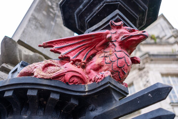 View of a vibrant red dragon statue perched atop a dark, ornate pillar, juxtaposed against the blurred backdrop of a grand building, Cardiff, Wales, United Kingdom.