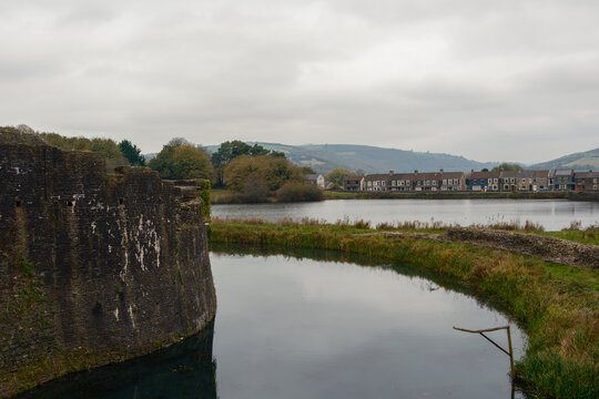 View of the ancient stone wall of Caerphilly Castle bordering the calm, reflective waters of the moat under a cloudy sky, Caerphilly, Wales, United Kingdom.