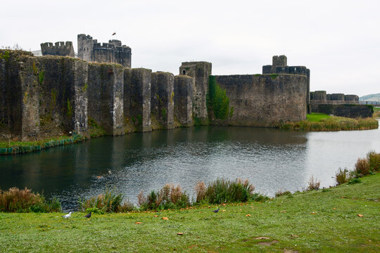 View of the imposing stone walls of Caerphilly Castle rising from the still moat waters under a brooding sky, a timeless fortress scene, Caerphilly, Wales, United Kingdom.