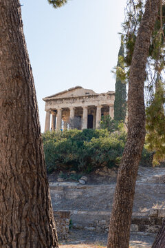 View of the ancient Temple of Hephaestus atop a verdant hill, framed by rustic tree trunks against a pale sky, evoking timeless beauty, Athens, Central Athens Regional Unit, Greece.