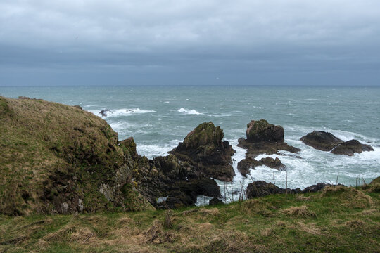 View of rugged cliffs meet the turbulent sea under a brooding sky, with the horizon blurred by mist, in a coastal scene, Peterhead, Scotland, United Kingdom.
