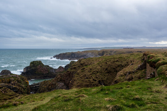 View of rugged cliffs meet the turbulent sea under a brooding sky, contrasting with patches of green grass, New Slains Castle, Peterhead, Scotland, United Kingdom.