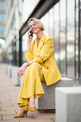 Fototapeta premium Full body smiling woman in yellow suit, sitting and talking with mobile phone in the city