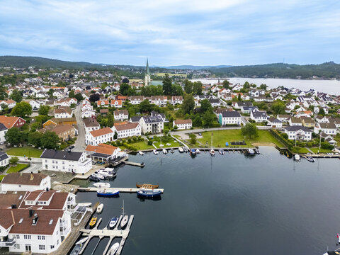 Aerial view of the quaint harbor where boats bob gently in the dark waters contrasting with the bright buildings, Lillesand Church rises in the background, Lillesand, Agder, Norway.