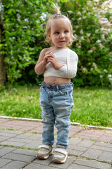 Resilient blond toddler with bandaged arm and charming top knot hairstyle, standing outdoors in jeans and sneakers on brick path against blurred backdrop of lush green leaves and white flowers.