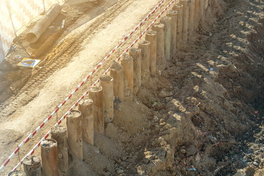 Overhead view of active construction site featuring sturdy retaining wall made of large concrete piles, bordering deep earth excavation with loose soil and dirt road protected by safety barrier under 