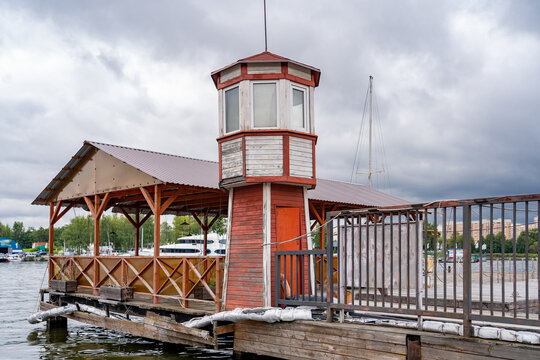 Weathered red and white wooden lookout tower and covered pier on cloudy day at marina with boats and distant city buildings