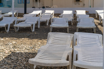 Numerous empty white plastic beach loungers standing ready on sun-drenched, pebbled shore, with coastal building in background, hinting at quiet relaxation.