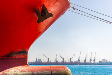 Middle East, Middle East - 18 December 2023: View of the bold red hull of a cargo ship, contrasted against the clear blue waters and distant port cranes, a maritime giant at rest.