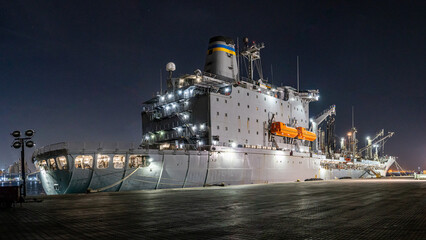 Middle East, Middle East - 18 December 2023: View of a colossal grey naval ship illuminated against the inky night, its structure casting long shadows on the slick, reflective ground.