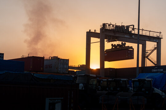 Middle East, Middle East - 19 December 2023: View of a colossal crane lifting a red container against the backdrop of a warm sunset, framed by rows of stacked containers and shadowed vehicles.