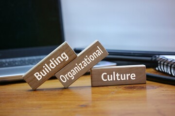 Wooden blocks with text 'building, organizational, culture' are placed on an office workspace alongside a laptop, notebook, and pen.