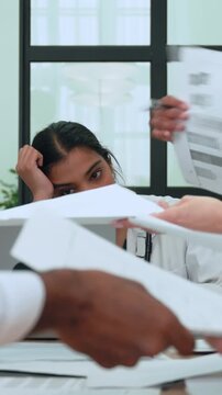 Medical professional overwhelmed by paperwork, colleagues adding more documents to her desk, illustrating the challenges of healthcare bureaucracy and administrative burden