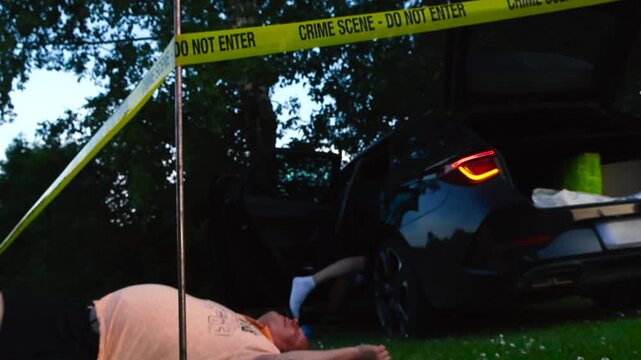 Low angle video of victims of a crime laying in and out of a crashed car with blinking lights while a yellow police lince Crime Scene tape is pulled around the area where the bloody victims are, night