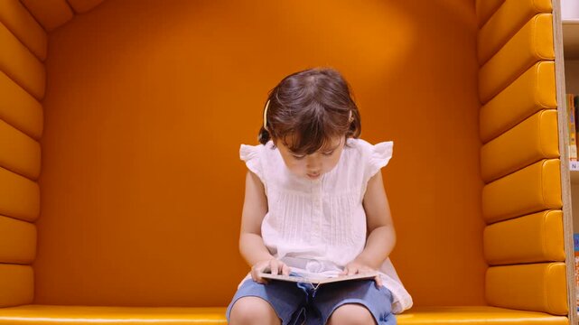 A cute 5 year old girl with yellow hairpins and pigtails sits in a small orange soft corner on an indoor playground, reading intently at a book.