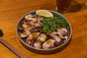 Thinly sliced octopus carpaccio served with tangy dressing, microgreens, and a lemon wedge on ceramic plate over wooden table in cozy dining setting
