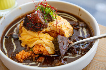 Japanese omurice with demi-glace sauce and braised beef chunks served in a white bowl, spoon revealing seasoned rice under soft omelet