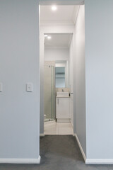 Narrow corridor leading to compact modern bathroom with tiled floor, glass shower, and white vanity sink, minimal residential interior