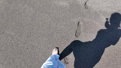 Shadow of a woman walking barefoot on a sandy beach, with visible footprints and bright sunlight highlighting the scene.
