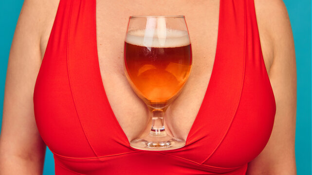 Close up of beer glass placed in red dress cleavage on blue background. Concept of Oktoberfest humor, playful lifestyle, beverage creativity, bold advertising, and festive cultural expression.