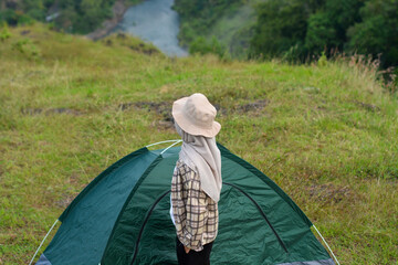 Sporty Muslim Asian woman standing near tent and backpack, enjoying sunny afternoon in the mountains. Camping lifestyle concept