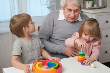 Obraz premium Grandfather playing toys with grandson and young granddaughter indoor. Old man grandpa studying with his grandchildren at weekend. Senior male having fun with kids. Happy multigeneration family video