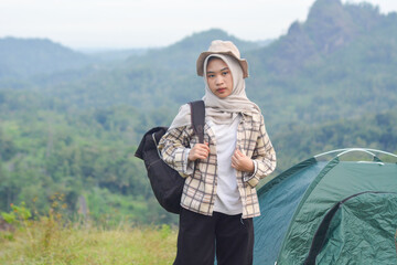 Sporty Muslim Asian woman standing near tent and backpack, enjoying sunny afternoon in the mountains. Camping lifestyle concept