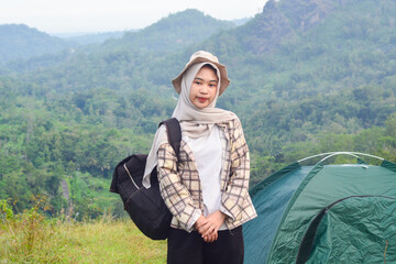 Sporty Muslim Asian woman standing near tent and backpack, enjoying sunny afternoon in the mountains. Camping lifestyle concept