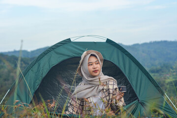 Sporty Muslim Asian woman standing near tent and backpack, enjoying sunny afternoon in the mountains. Camping lifestyle concept