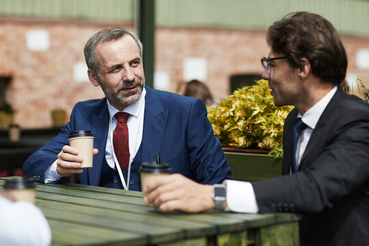 Middle aged Caucasian man with gray hair and beard talking to young businessman with brown hair, while holding coffee cups, outdoors at wooden table during meeting - Powered by Adobe