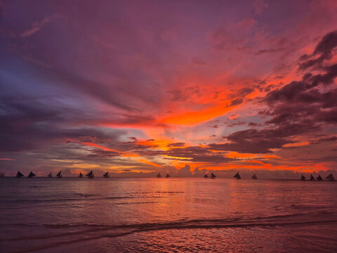Spectacular sunset seascape at Boracay White Beach Philippines with traditional paraw sailboats silhouetted on the horizon, vibrant orange purple and pink sky reflecting on calm tropical ocean water