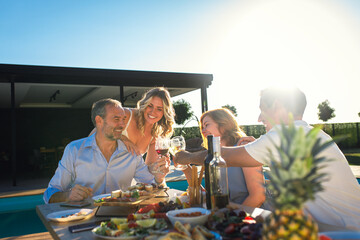 Group of adult friends having lunch by swimming pool in sunny backyard and toasting with wine.