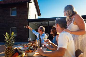 Group of adult friends having lunch by swimming pool in sunny backyard making selfie.