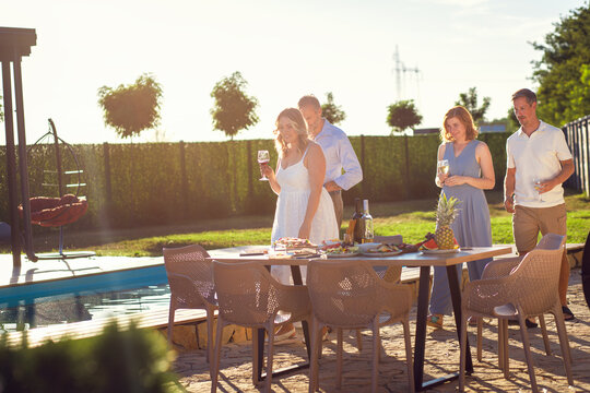 Group of adult friends talking by swimming pool drinking wine in sunny backyard.