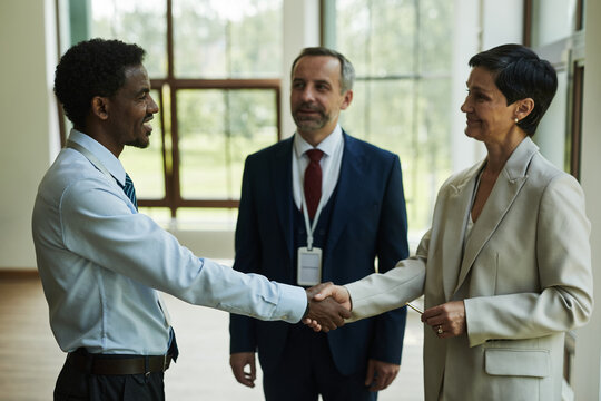 Caucasian middle aged woman and Black man shaking hands in office hallway, while Caucasian colleague is observing interaction, all dressed in business attire, diverse business partners making deal