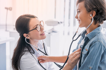Female pediatrician examining boy in modern medical clinic using stethoscope, listening to breath, heartbeat, pneumonia symptoms in lungs. Nurse medical worker checking up on child kid in hospital