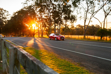 Panning image of red car driving past on sealed rural country road in sunset light