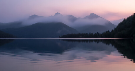 Fototapeta premium A calm mountain lake reflects a foggy landscape during a dusky sunrise.