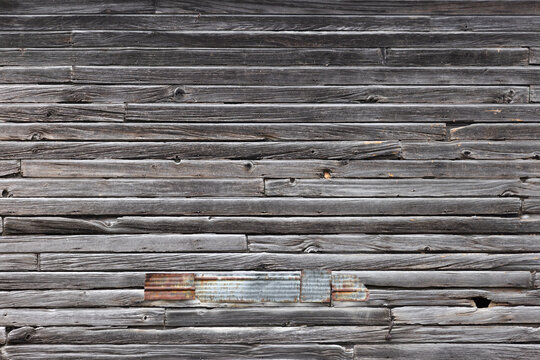 Wall of old weathered horizontal wooden planks with a section patched or repaired with pieces of corrugated metal adding to the rustic and utilitarian aesthetic - Powered by Adobe