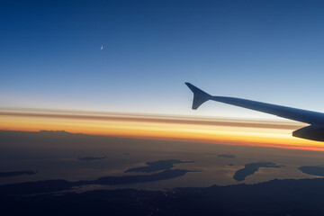 Fototapeta premium Stunning aerial view of a sunset over islands, seen from an airplane window. A crescent moon is visible in the clear sky.