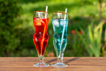 Two artistic glasses shaped like a nude woman and man filled with red and blue alcoholic cocktails on table with lush green garden background, closeup. Playful summer vibe for creative drink concepts