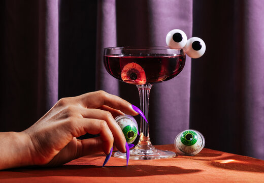 Female witch hand with fake nails holding eyeball by purple cocktail
