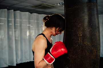 Exhausted female boxer, wearing red gloves, resting her head against a black heavy bag after an intense workout in a boxing gym, showcasing the toll of her rigorous training