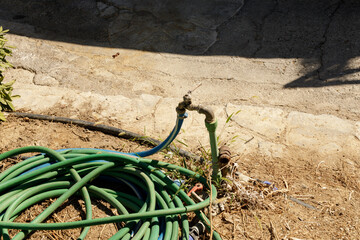 Green garden hose coiled near a weathered outdoor faucet. Dry, cracked ground. Summer.
