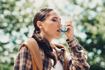 Young woman using an asthma inhaler outdoors on a sunny day surrounded by nature emphasizing health...