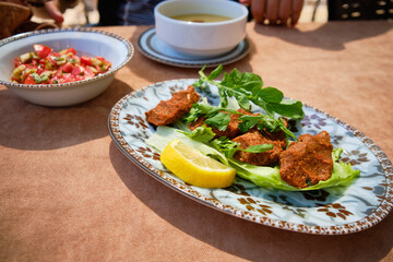 Close-up view of traditional Turkish dish called cig kofte served on bed of crisp lettuce, garnished with fresh parsley and lemon slices. Bowls of tomato salad and lentil soup at background