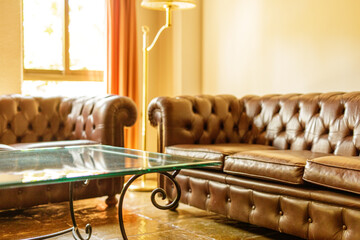 Elegant brown leather chesterfield sofa and glass coffee table in a sunlit living room. Perfect for...