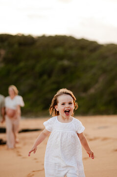 Young girl in white dress running on sandy beach at sunset while family walks behind her
